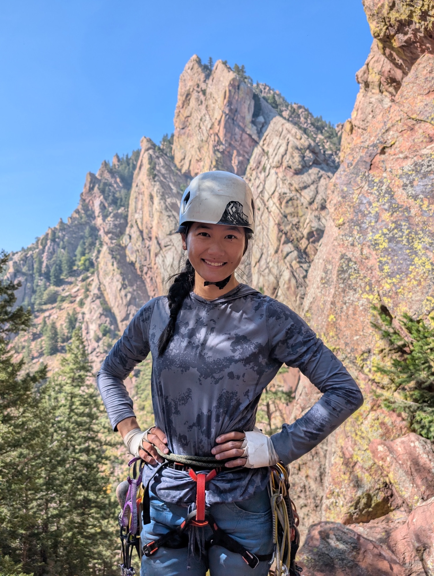 Mia standing, looking at camera and smiling wearing a helmet, harness and climbing gear in Eldorado Canyon. Redgarden wall (big rock cliff) is in the background.